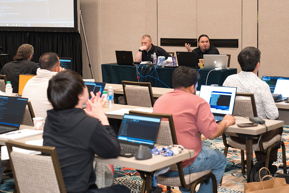Two instructors with laptops sit at a table before rows of students, also seated at tables with laptops in front of them. One instructor is gesturing as he talks to his students.
