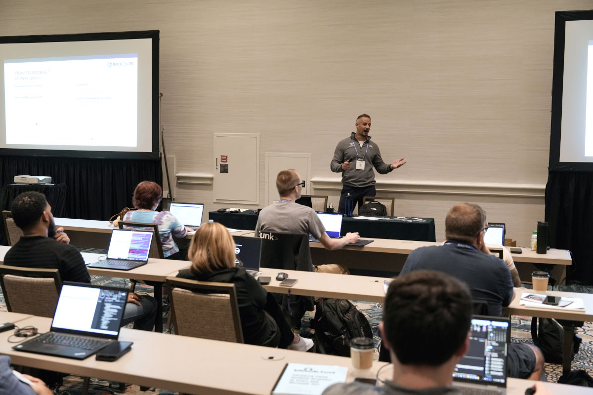 One instructor stands between two presentation screens and gestures as he speaks to his students. The students are seated in rows facing the instructor with their laptops open.